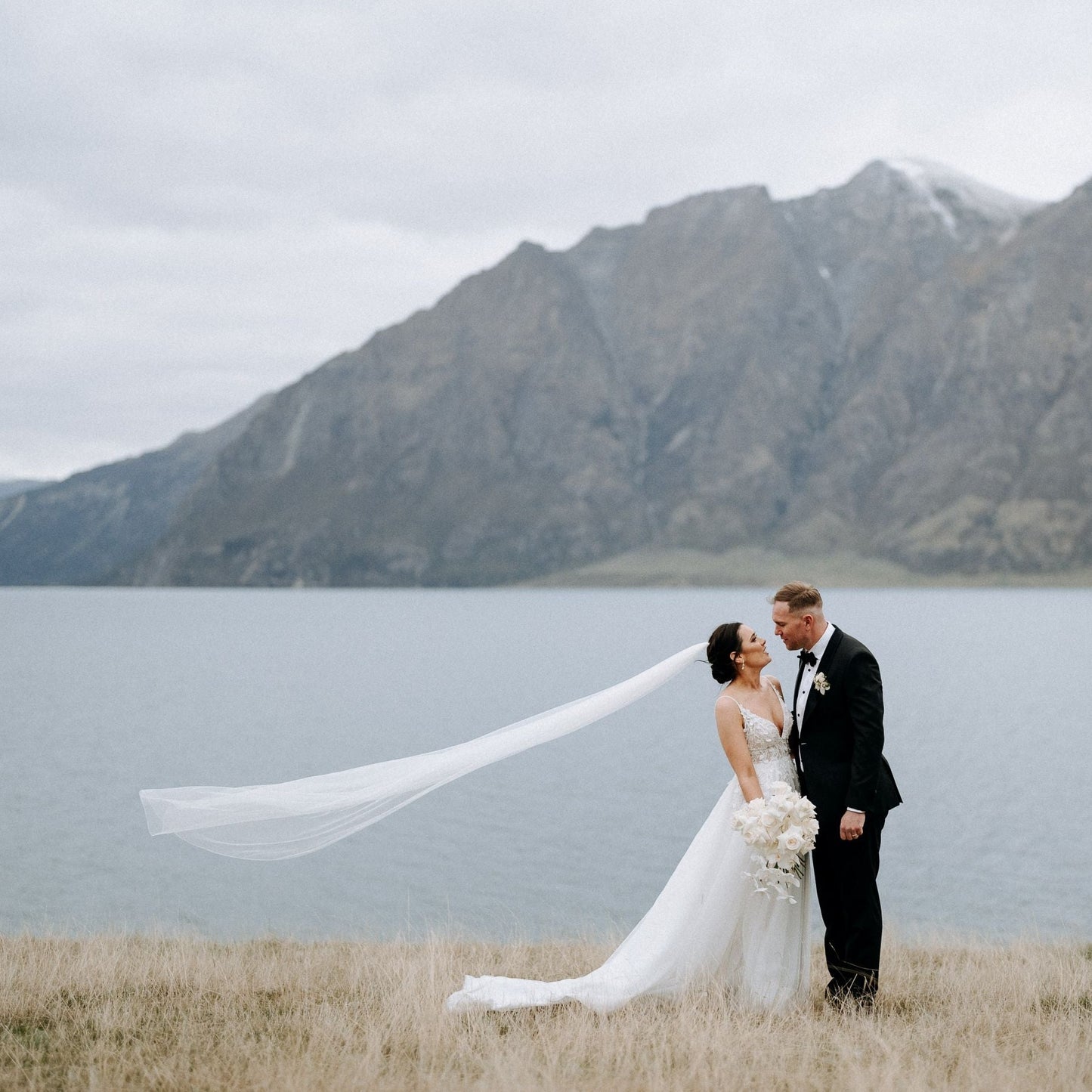 Bride and groom stand in front of lake and mountain, veil flows in wind