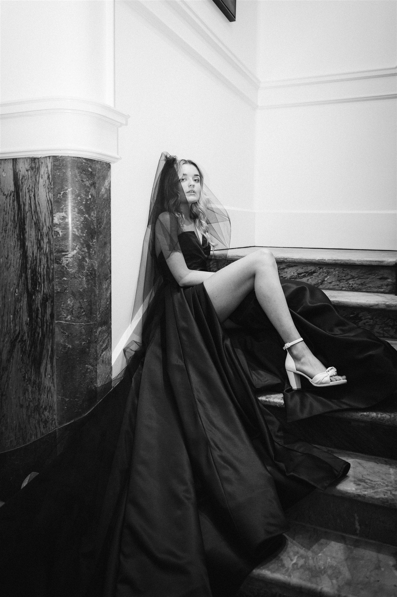Model sits on marble stairs wearing black Daisha wedding dress with dramatic split.