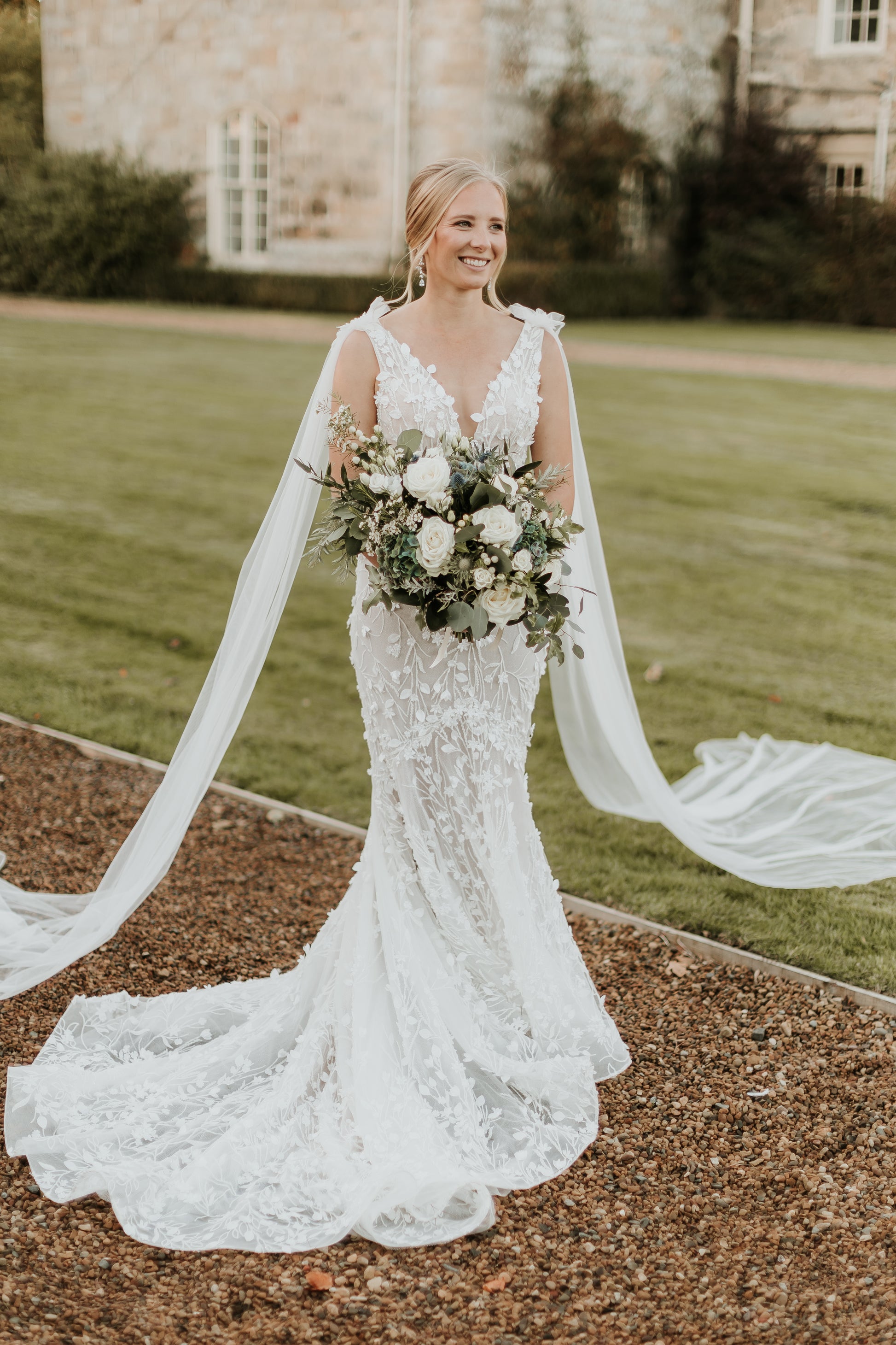 Bride wears Saige wedding gown. A mermaid cut dress with long tulle wings. She holds a large bouquet.