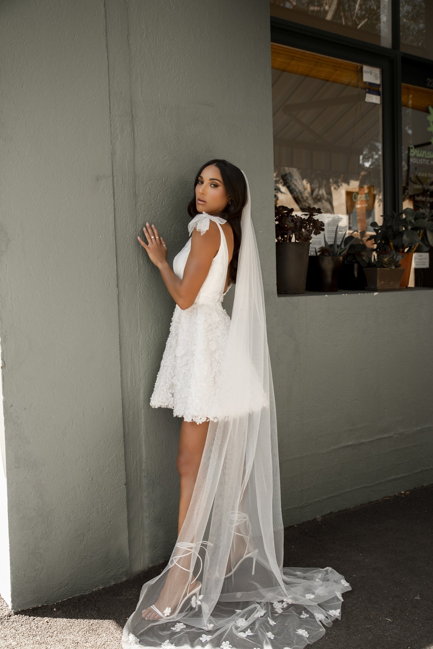 Bride stands against wall in white mini dress with long 3D floral veil.