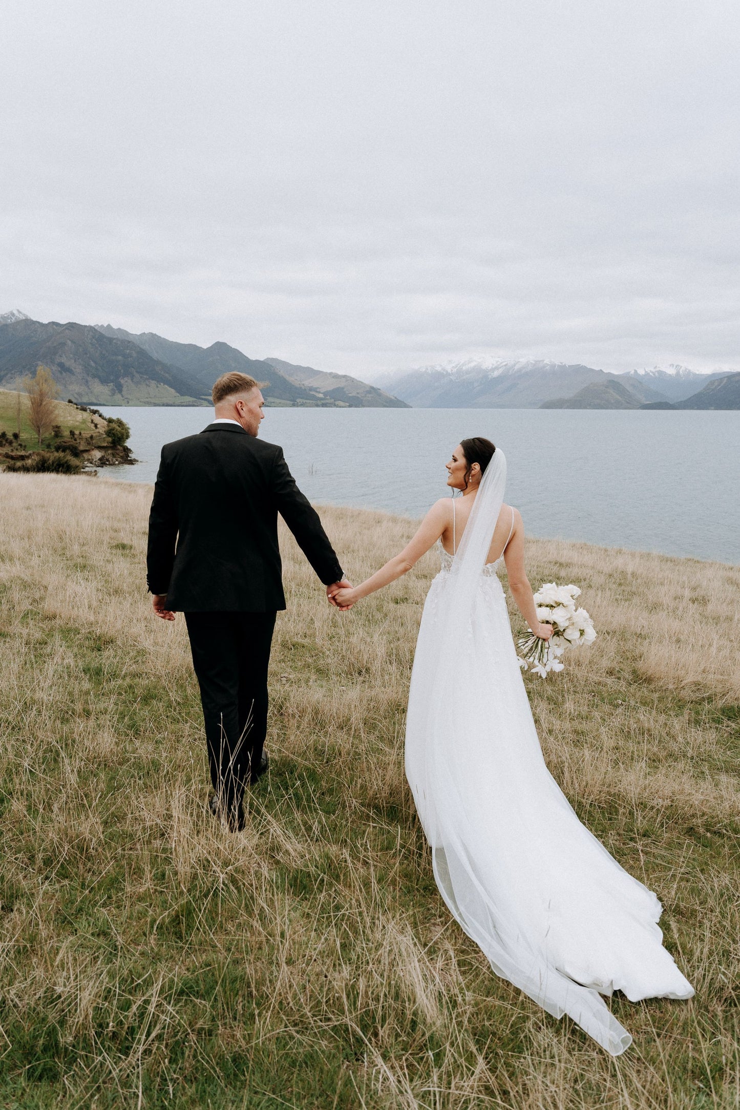 Bride and groom hold hands near lake, three metre veil trails behind.