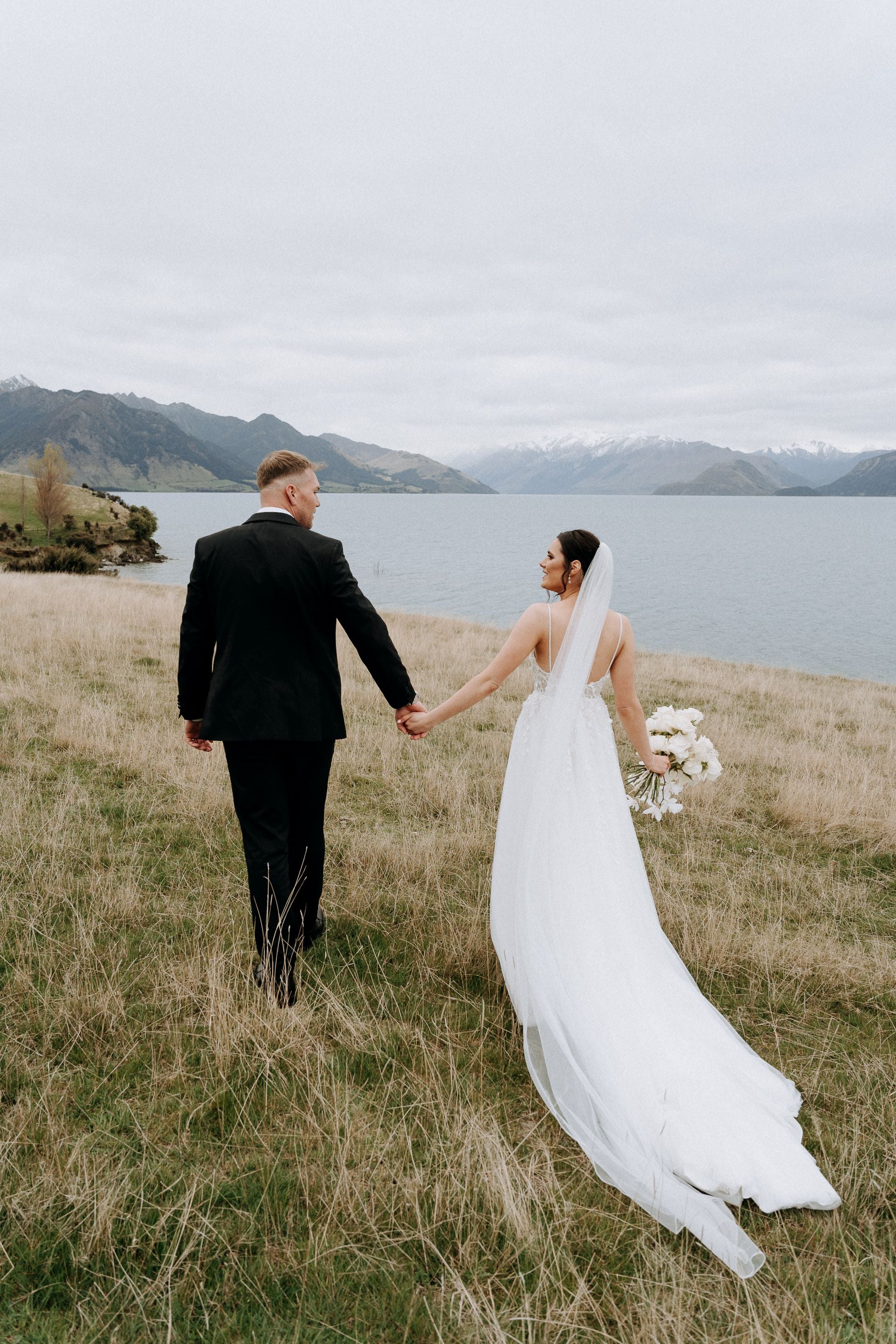 Bride and groom hold hands near lake, three metre veil trails behind.