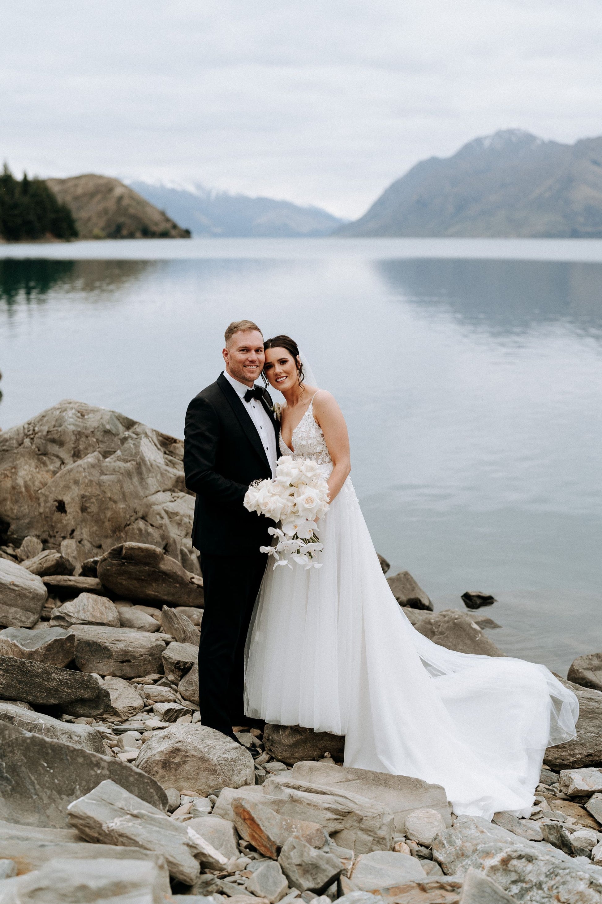 Bride and groom smile in front of lake and mountains.