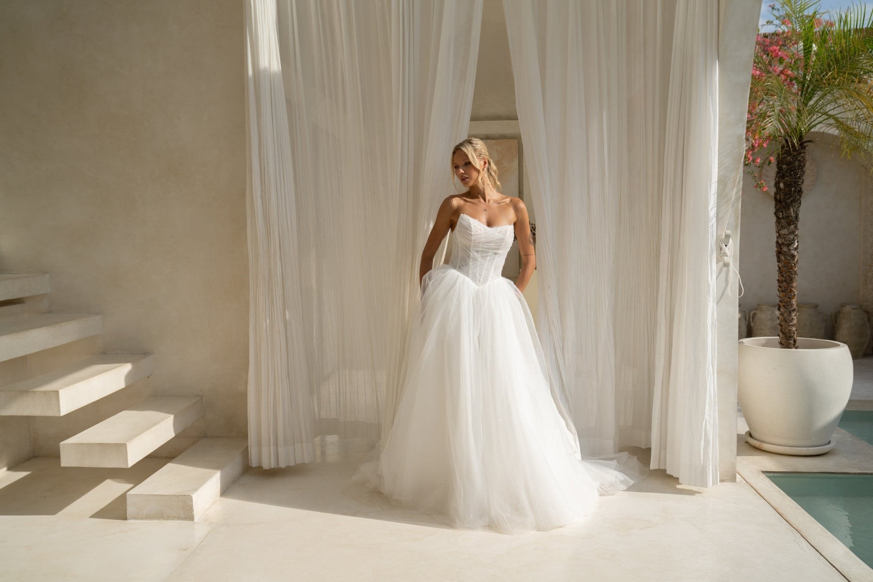 A-line wedding gown with cat-eye strapless neckline, criss cross ruching around the bust, corset boning on the bodice, basque waist and a voluminous tulle skirt. 