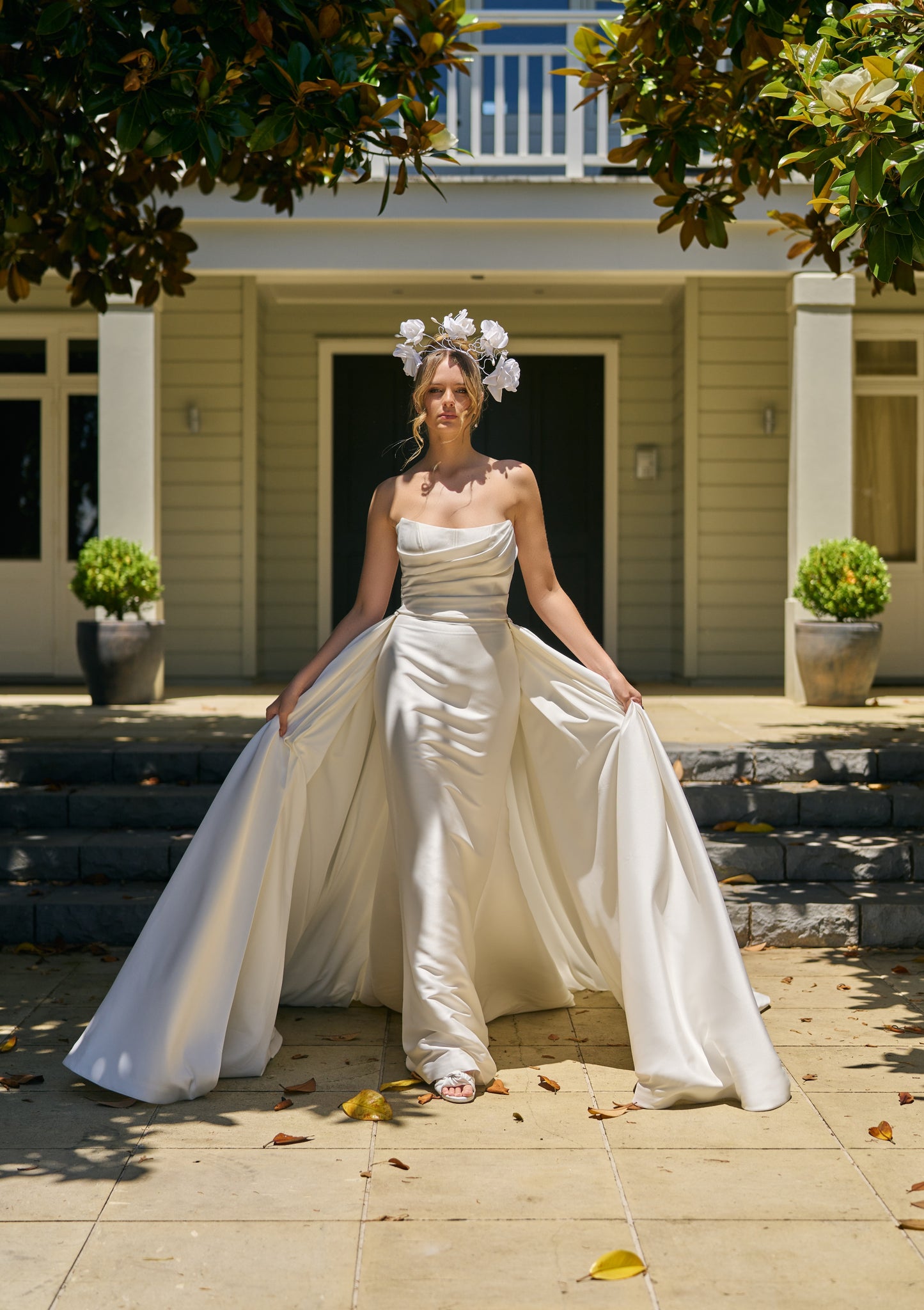 Model walks with Lisha gown and detachable Lisha overskirt that trails behind her from the side seams. Gown is strapless and ruched through bodice