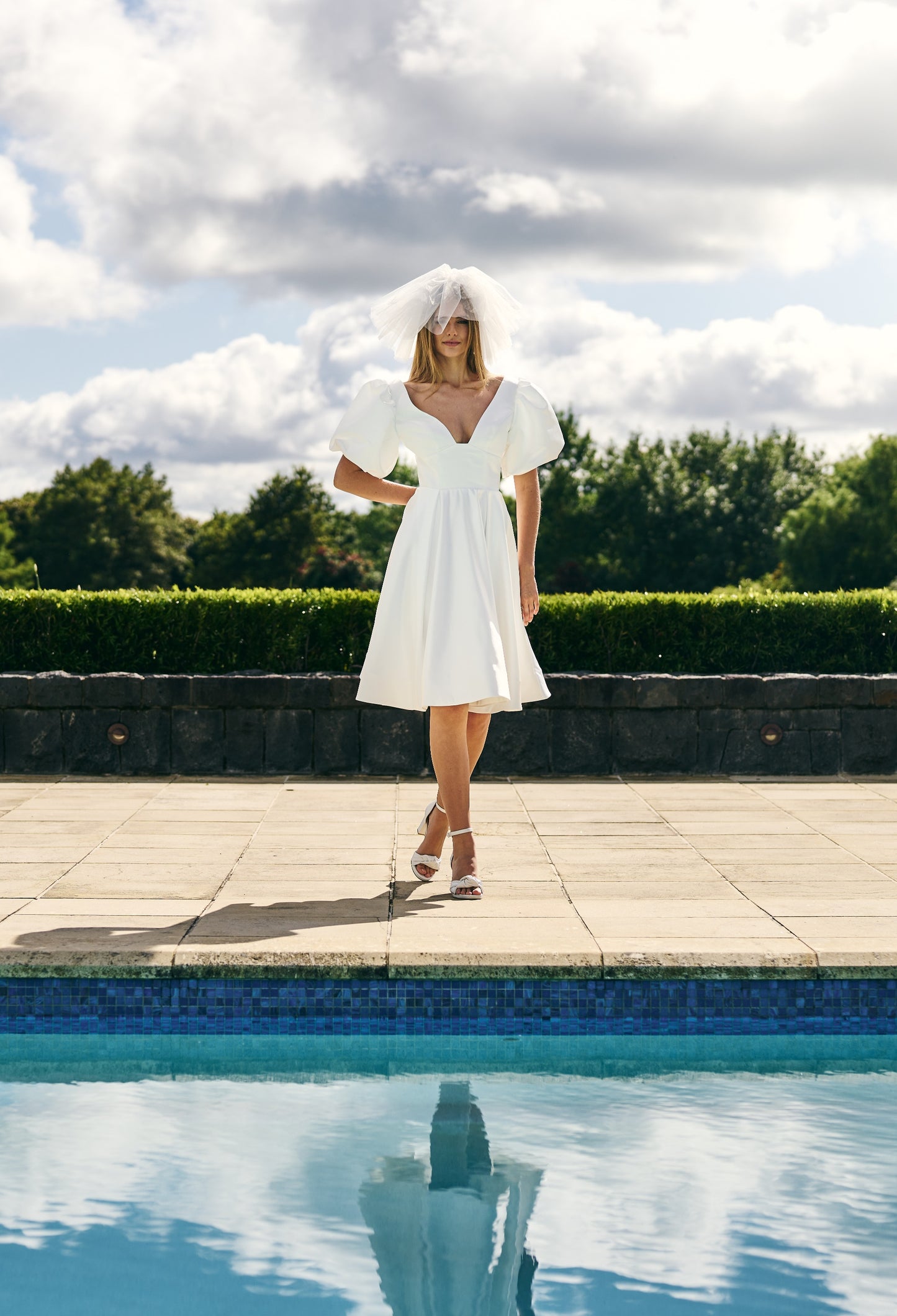 Bride stands in front of pool in mini dress and short fascinator-like veil