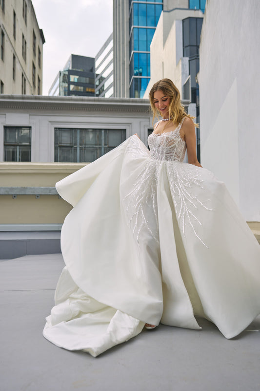 Raffael wedding dress from Jessica Coutures. Square neckline, low back A-line dress with organza fabric and glitter detailing at the bodice. Boning support, cinched waist, dramatic skirt. Hidden features include pockets and a split for easy movement.