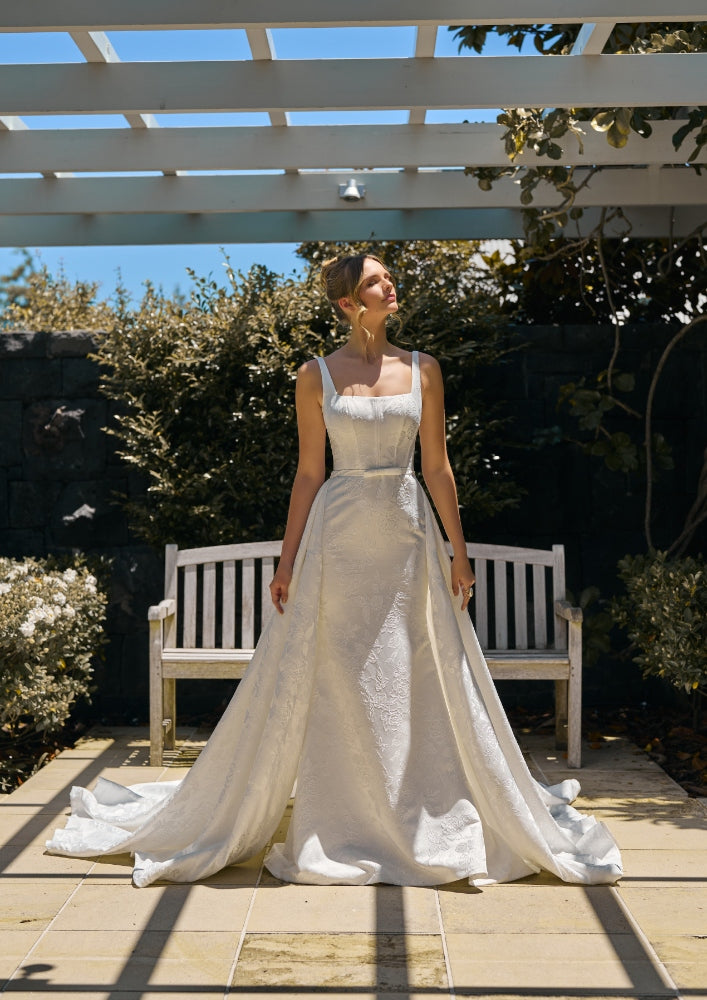 Model wears Perry dress with detachable perry overskirt in ivory jacquard fabric. 